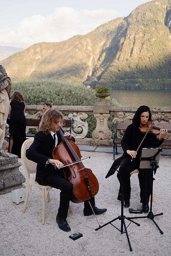 Wedding musicians in black formal attire play violin and cello at a stone terrace ceremony, with lake and mountains beyond the balustrade