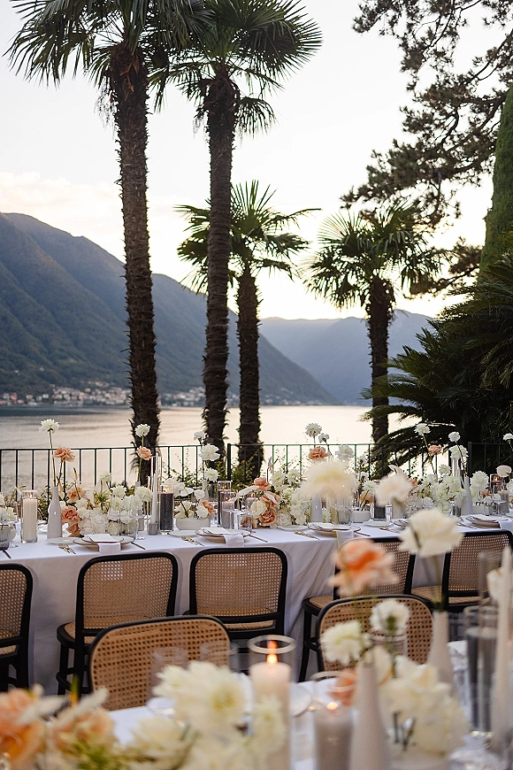 Reception tablescape with white tablecloth, low white and peach flowers, bud vases, and pillar candles on a terrace with lake and mountains beyond