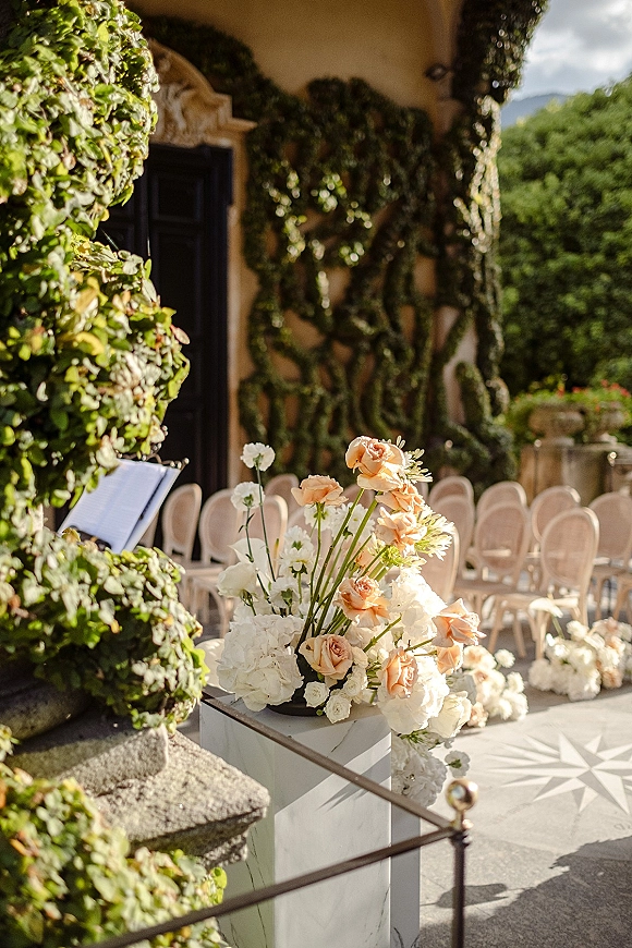 Ceremony aisle decor with aisle floral arrangements of peach roses and white hydrangeas on plinths beside chairs on a stone terrace
