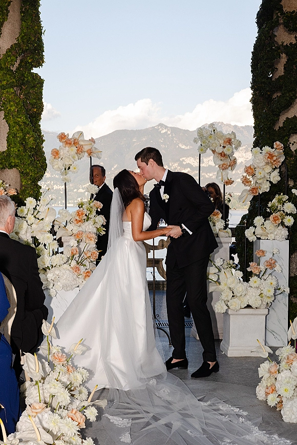 Ceremony kiss as bride and groom embrace beneath a floral arch with white and peach blooms on an outdoor terrace by lake and mountains