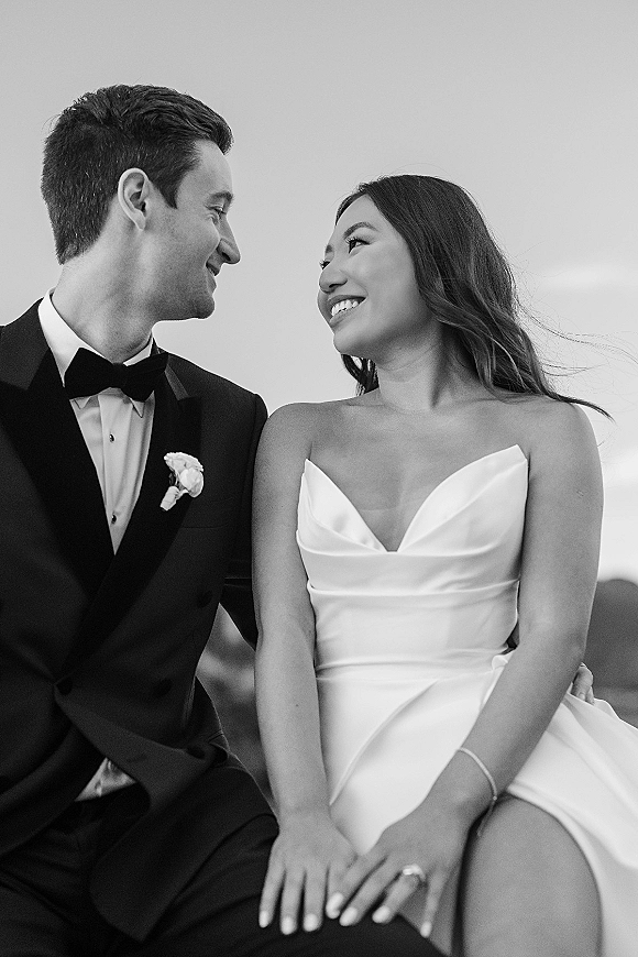 Couple portrait in a black and white wedding portrait style, bride and groom seated holding hands under open sky with distant hills
