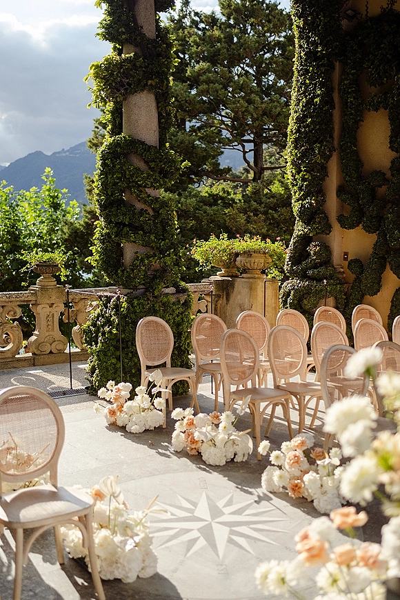 Ceremony setup with outdoor ceremony chairs, cane bistro seating and low white and peach floral clusters on a stone terrace with mountain view