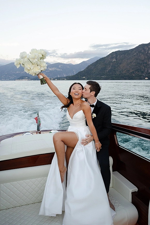 Wedding couple portrait sharing a wedding kiss moment on a wooden boat, bride holding a white rose bouquet with mountains and lake wake behind