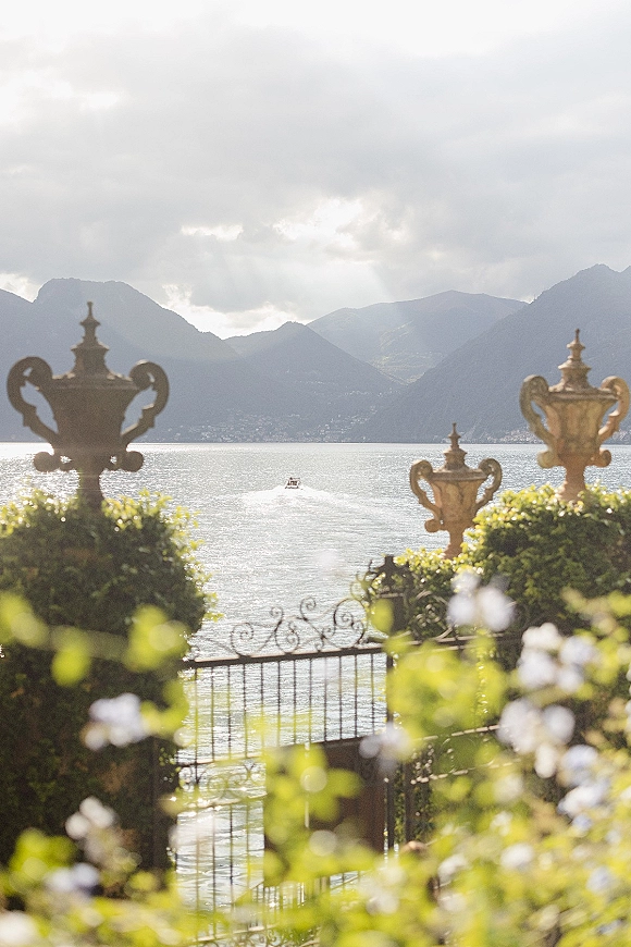 Wedding venue view with a lakefront wedding venue terrace framed by wrought iron gate, urn planters, and hedges, overlooking lake and mountains