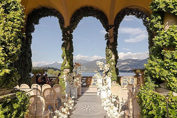 Ceremony setup with outdoor ceremony arch framed by orchids and white-peach aisle florals on a terrace overlooking lake and mountains