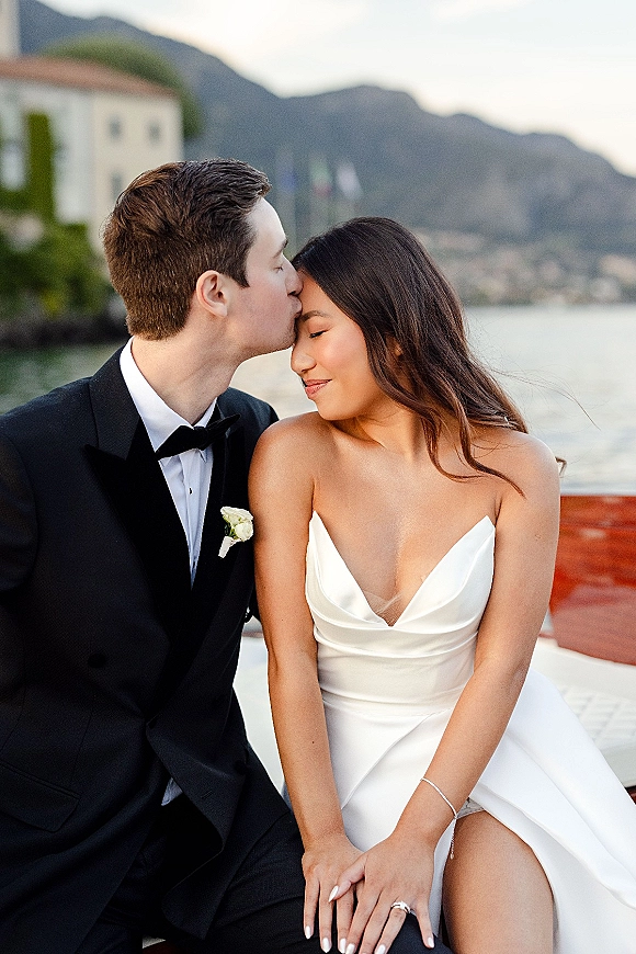 Couple portrait of bride and groom kiss on a wooden boat, her strapless satin dress and his black tuxedo against a mountain lake backdrop