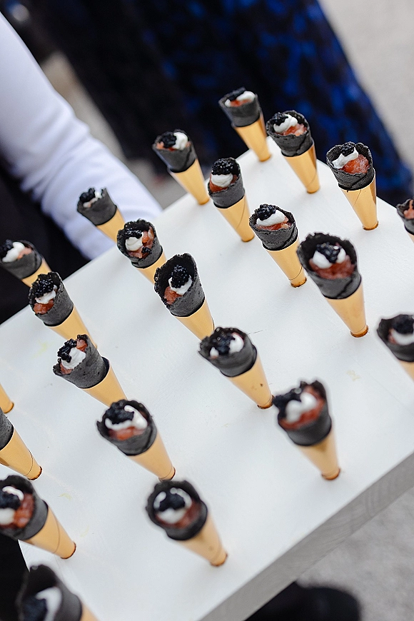 Wedding appetizers on a serving board of black mini cones filled with savory cream and caviar, carried by a server outdoors with guests blurred behind