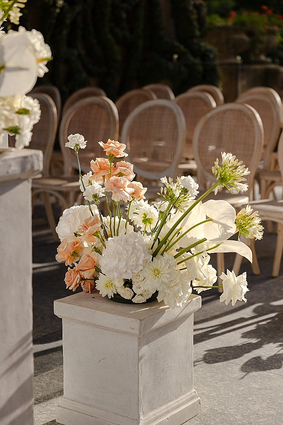Ceremony aisle decor with an aisle floral arrangement of peach roses, white blooms and greenery on a stone pedestal in a courtyard walkway