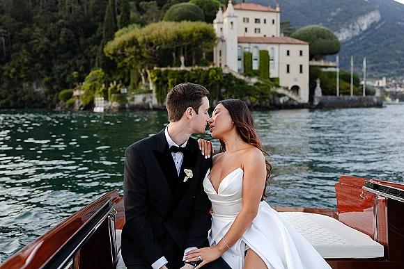 Wedding kiss portrait of bride and groom on boat, her strapless satin dress and ring visible, with lake, villa, and mountains behind