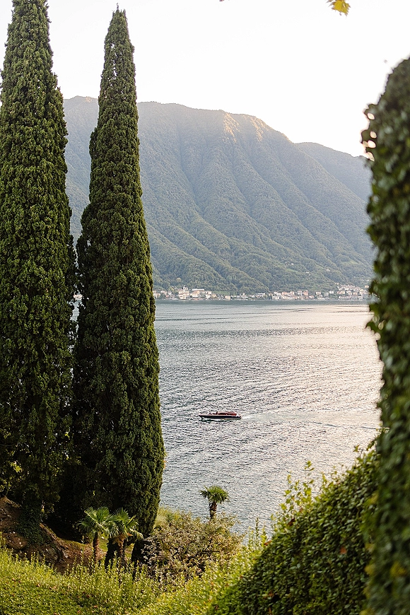 Lake landscape with cypress trees by lake, framed by palm and shrubs, overlooking a mountain shoreline village and a small motorboat on water