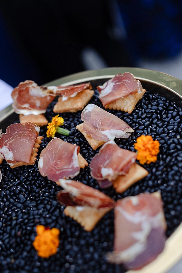 Wedding cocktail appetizers of passed hors d'oeuvres with prosciutto on crackers atop black lentils, marigold garnish on a tray in blurred reception