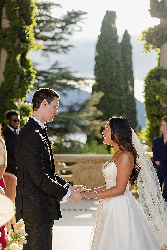 Wedding vows as bride and groom hold hands on an outdoor terrace, her long veil flowing by a stone balustrade with lake and mountains behind