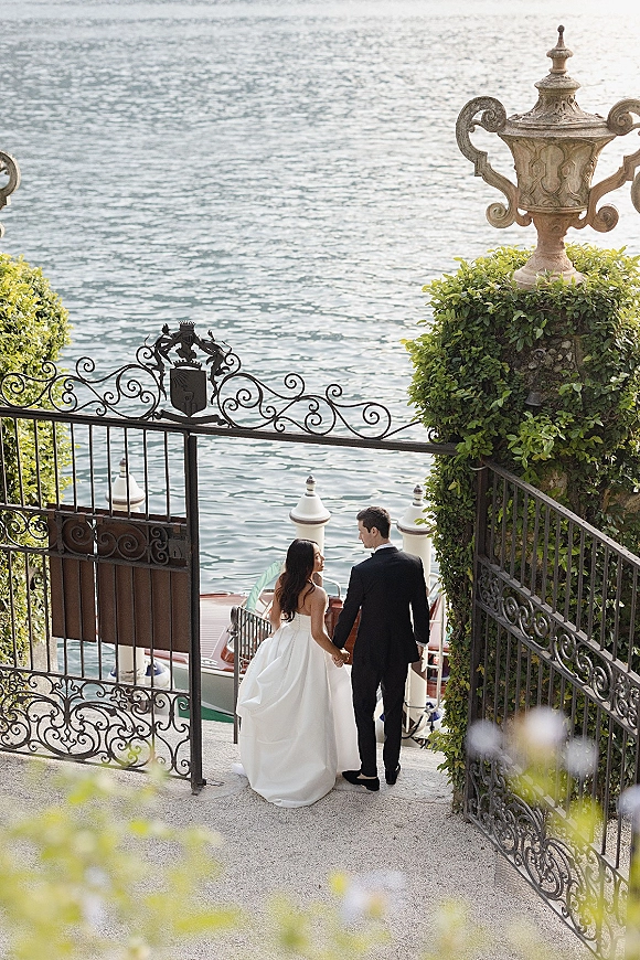 Couple portrait of bride and groom holding hands from behind, walking down stone stairs by wrought iron gate toward lake dock and boat