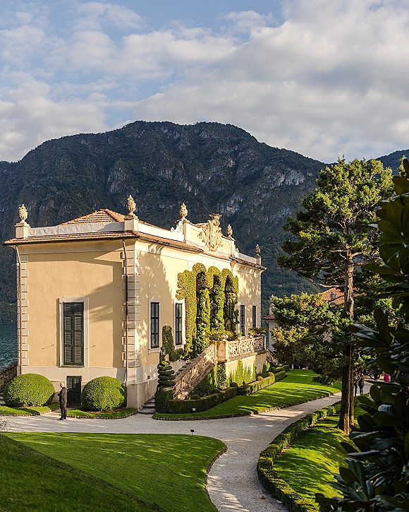Wedding venue exterior with a historic villa, stone staircase and terrace balustrade, framed by topiary gardens and lake mountains beyond