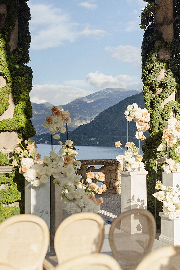 Ceremony setup with outdoor ceremony decor, white and blush floral arrangements on plinths lining an aisle on a terrace overlooking lake and mountains