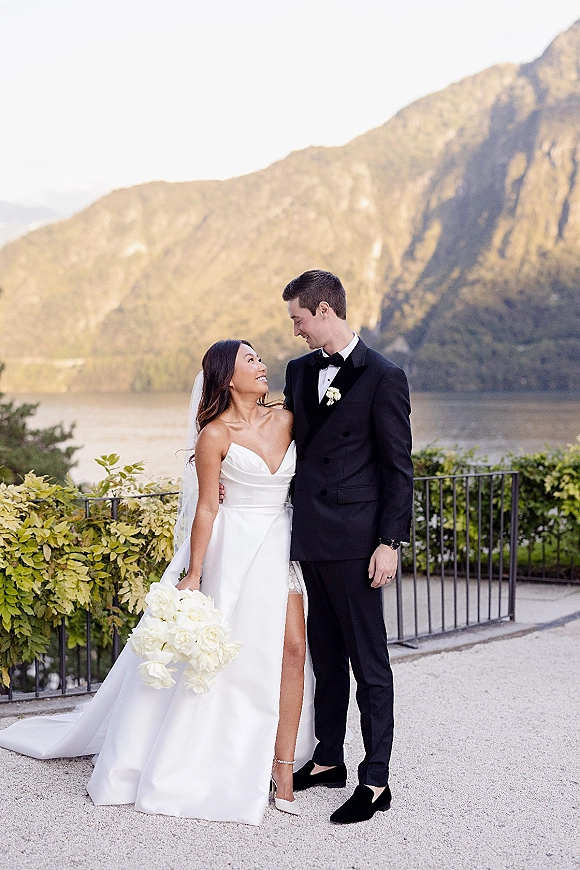 Couple portrait of bride and groom looking at each other, bride holding white rose bouquet on terrace with lake and mountains behind