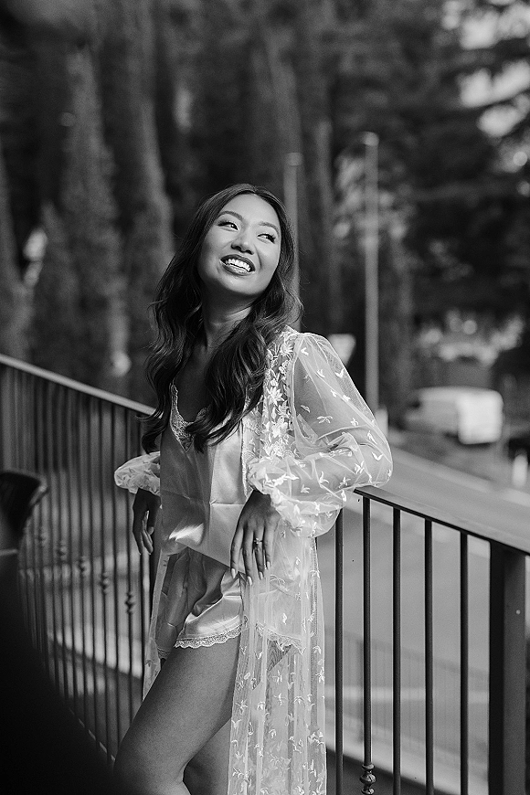 Bridal portrait of a bride in a lace robe over lingerie, leaning on a balcony railing with trees and a blurred street behind