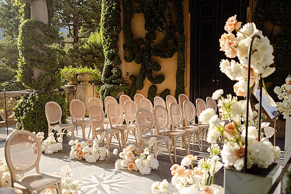 Ceremony setup with outdoor ceremony seating, cane-back chairs and white-blush aisle flowers on a sunlit stone terrace by ivy walls