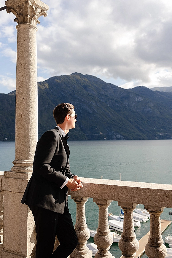 Groom portrait in a black tuxedo and sunglasses, leaning on a stone balcony with lake, boats, and mountains under cloudy sky