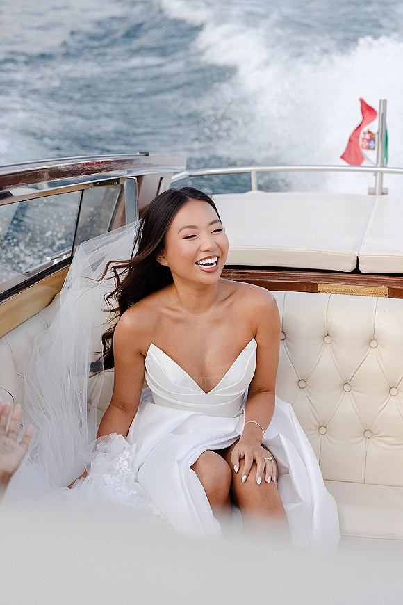 Bridal portrait of a bride on boat laughing in a strapless satin gown, veil blowing over tufted seats with ocean wake behind