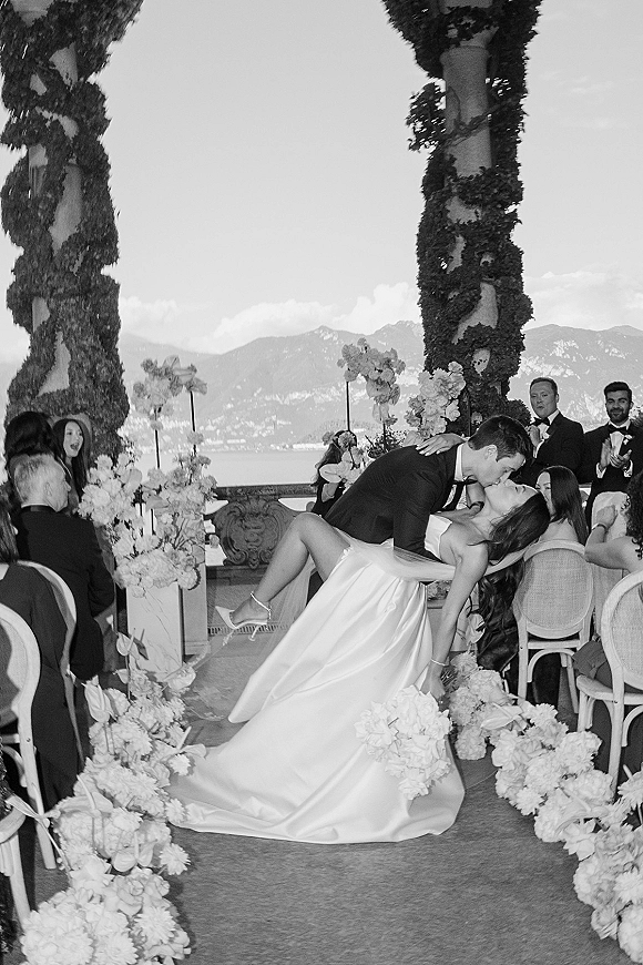 Wedding kiss portrait as groom dips the bride in veil and gown, with floral-lined aisle on a terrace overlooking lake and mountains