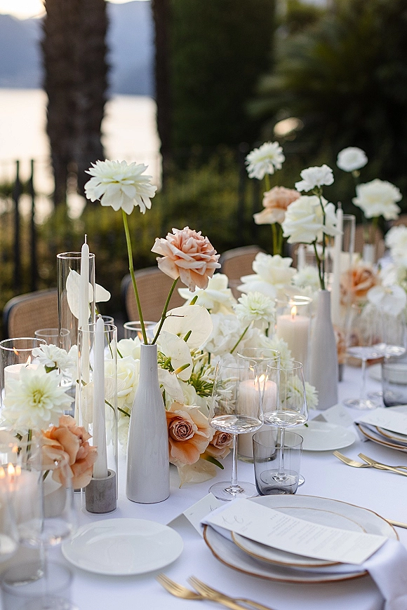 Reception tablescape with wedding table centerpiece of white and peach flowers, taper candles, gold flatware, on an outdoor patio by water view