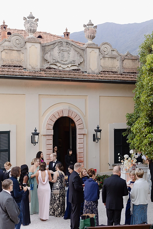 Wedding cocktail hour with wedding guests mingling by a bar table of drinks, floral accent, and lantern-lit villa courtyard with mountains