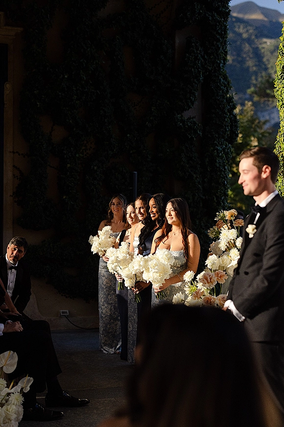 Ceremony moment with wedding ceremony party as bridesmaids hold white and blush bouquets beside a groom in black tux, ivy wall and mountains behind