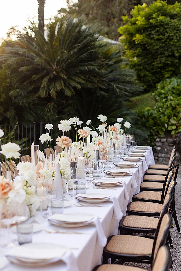 Reception tablescape on a long banquet table with blush and white bud vases, pillar and taper candles, rattan chairs in a garden patio setting