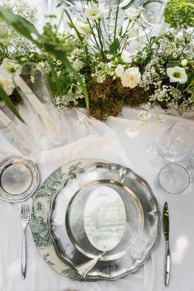 Reception tablescape with wedding table centerpiece of white florals and greenery on a moss runner, with taper candles, crystal glassware, and silver chargers