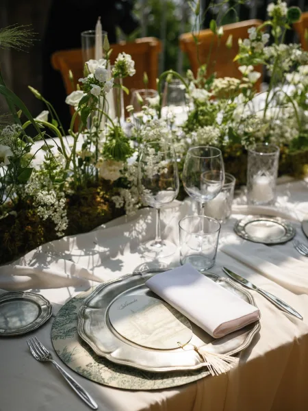 Reception tablescape with wedding table centerpiece of white flowers, moss and greenery garland, taper candle, and silver chargers in sunlit garden setting