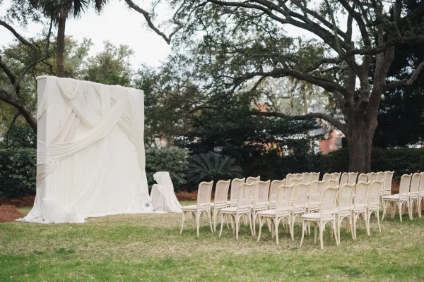 Ceremony setup for an outdoor wedding ceremony with a white draped fabric backdrop, aisle runner, and chairs on a grassy lawn beneath oak trees