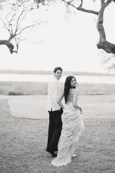 Couple portrait in black and white, bride and groom walking away then looking back, her lace open-back gown and heels by a lakeside field