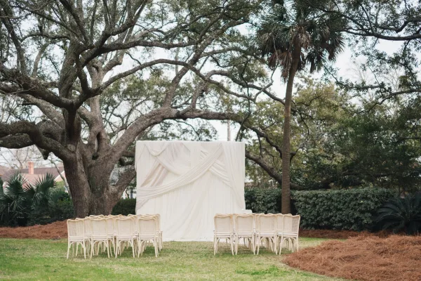 Ceremony backdrop with draped fabric wedding backdrop in white, framed by oak trees and lawn, with rows of white chairs facing it