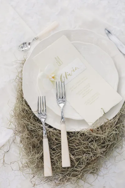 Wedding place setting with layered white plates, monogram menu and calligraphy name card, ivory flatware, and white orchid on moss runner