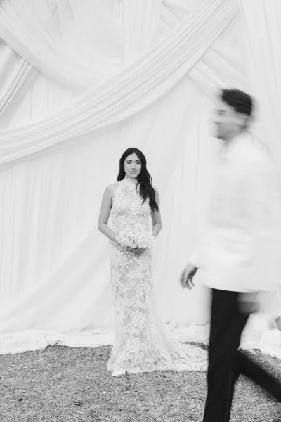 Bridal portrait of a bride holding a bouquet in a lace wedding dress with a high neck gown before white curtain drapery on grass