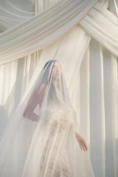 Bridal portrait of a bride with veil over her face, wearing a lace high-neck wedding dress and holding a white bouquet before white drapery