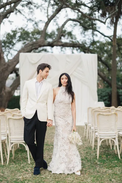 Couple portrait of bride and groom holding hands, bride in lace gown with white bouquet, walking past chairs on a lawn with trees