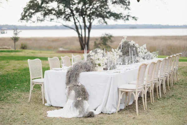Reception tablescape with an outdoor wedding reception table featuring white linens, cane chairs, taper candles, and greenery by the water