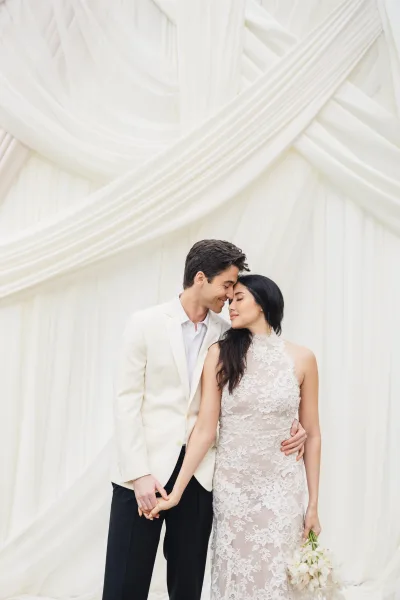 Couple portrait of bride and groom holding hands, touching foreheads, in lace gown and white jacket with lily bouquet before draped backdrop