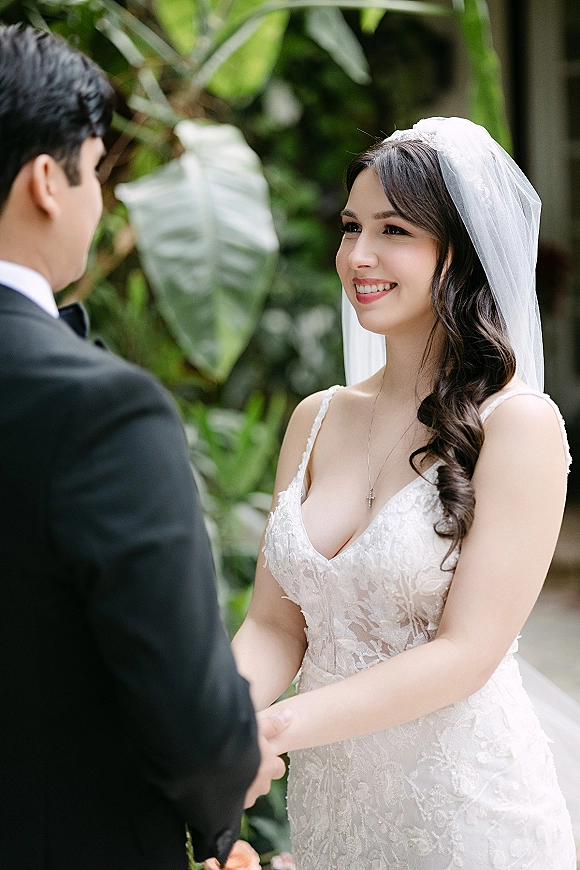 Wedding vows as bride smiles at groom, holding hands in lace dress and veil beside tuxedoed groom against tropical greenery backdrop