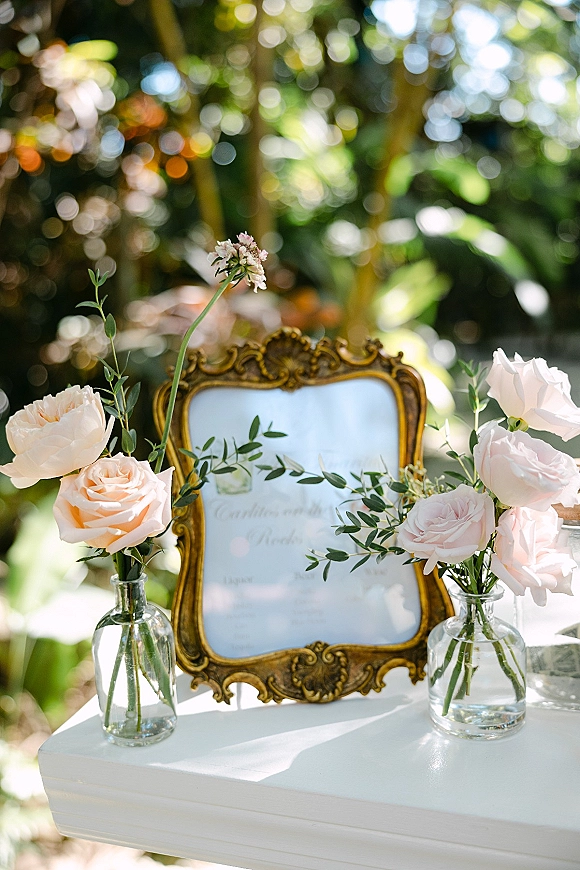 Wedding welcome sign on a mirror with ornate gold frame, blush roses and greenery, on a white pedestal amid sunlit garden foliage bokeh