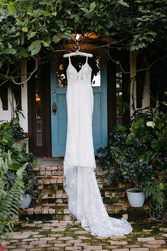 Wedding dress hanging on a front door, lace wedding dress with spaghetti straps and V neckline, train draped over brick steps with greenery