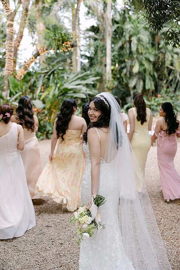 Bride portrait with bride looking back in a lace dress and long veil, holding a blush bouquet on a gravel path in a tropical garden