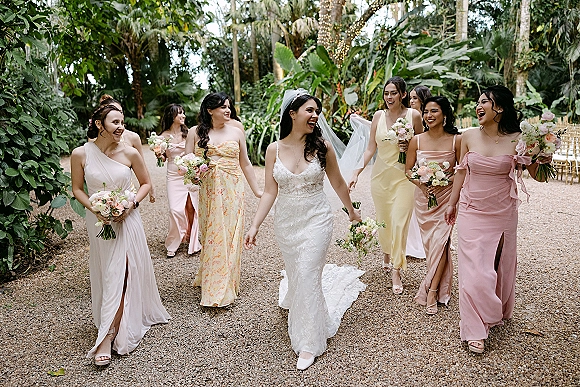 Bride with bridesmaids walking with bride along a gravel path in a tropical garden, holding bouquets under string lights and palm trees