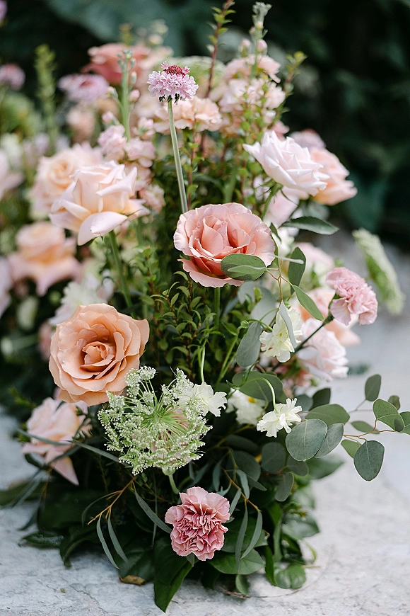 Wedding floral arrangement of peach rose wedding flowers with pink carnations and eucalyptus on a stone surface, soft garden foliage behind