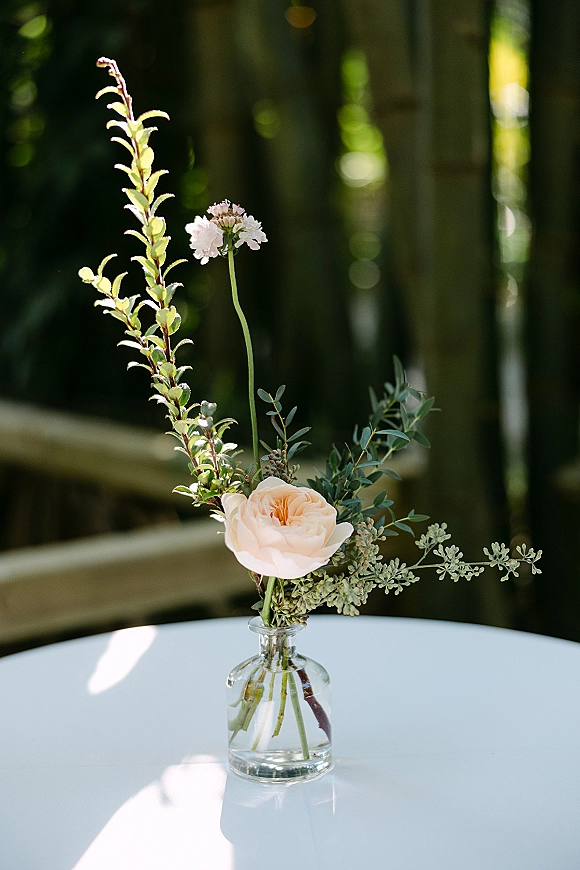 Bud vase centerpiece with a peach rose and greenery in a clear glass vase on a white round table, sunlit with blurred trees behind