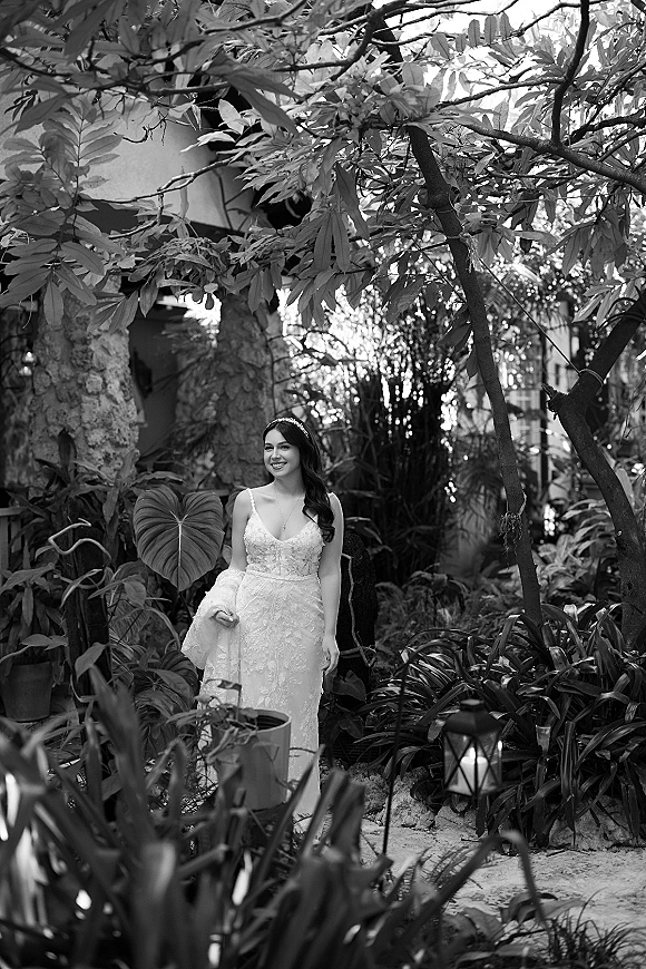 Bridal portrait of a bride in a lace wedding dress, smiling as she holds her train on a garden walkway near a lantern and stone wall