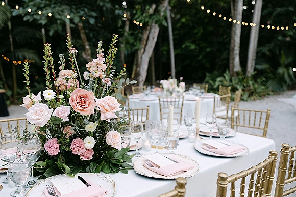 Reception tablescape with a wedding table centerpiece of blush roses and greenery on white linens, gold chiavari chairs under string lights outdoors