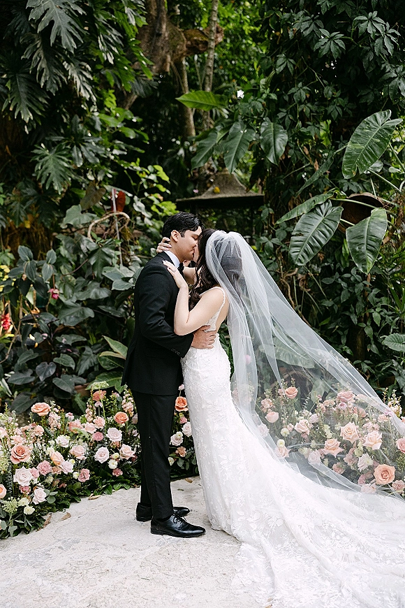 Wedding kiss portrait of bride and groom kissing, her long veil and lace dress flowing as they embrace on a stone patio amid tropical greenery